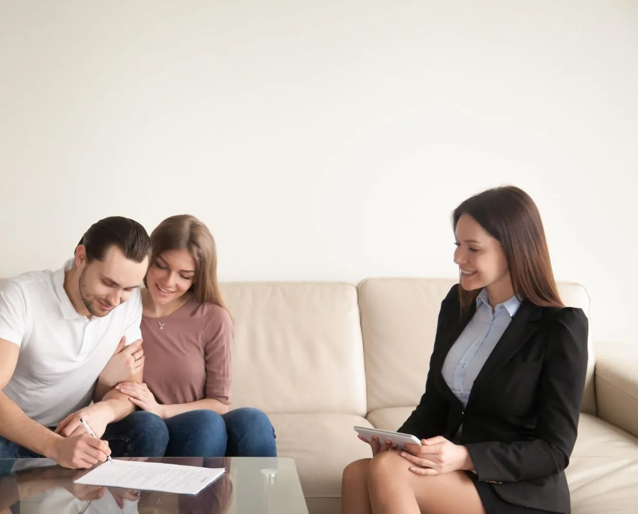 Couple signing document, real estate agent smiles. Living room setting; couple on couch, agent in suit.