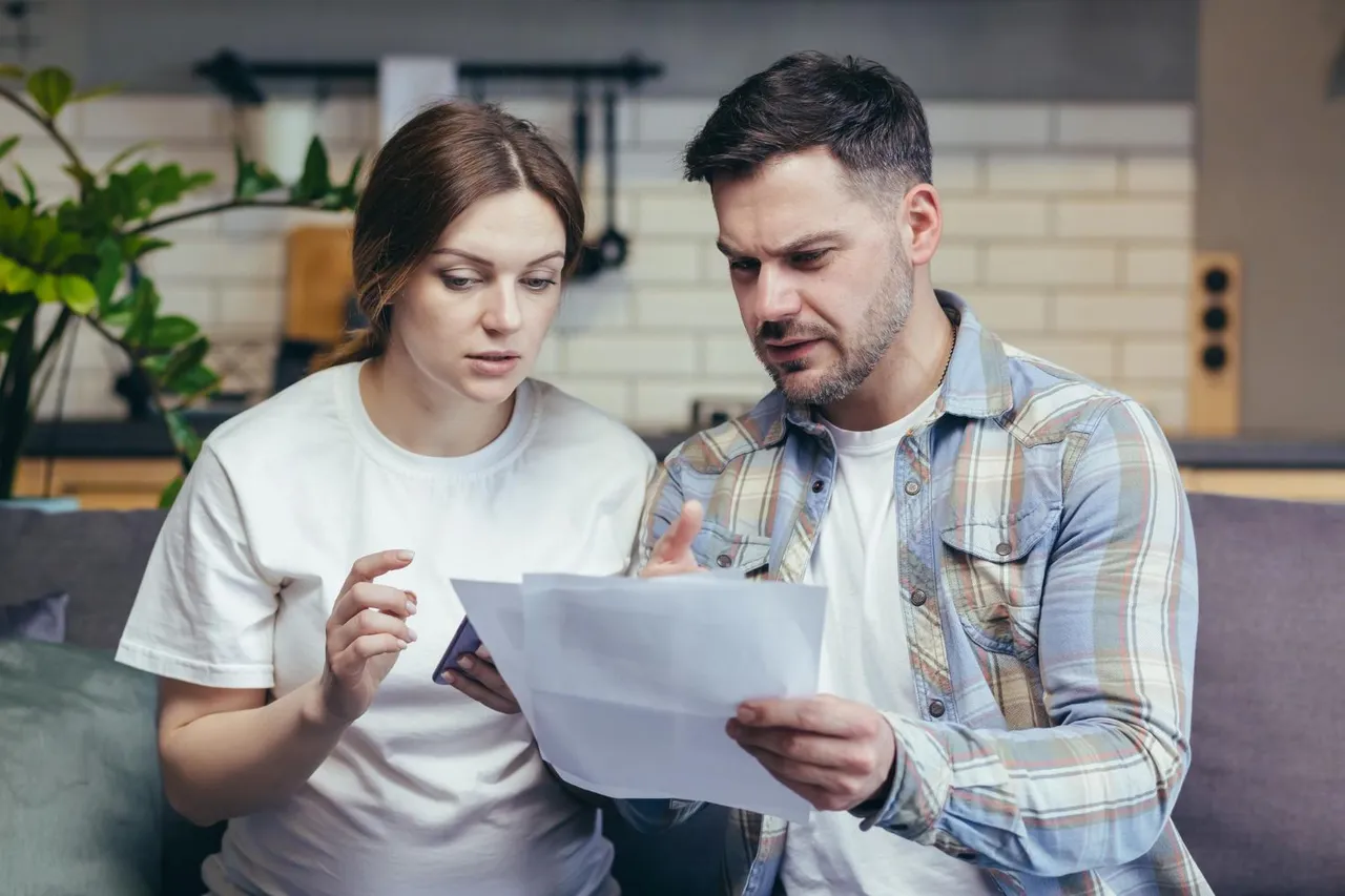 Couple looking at documents, appearing concerned, in a living room setting.