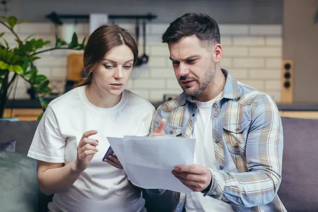 Couple looking at documents, appearing concerned, in a living room setting.