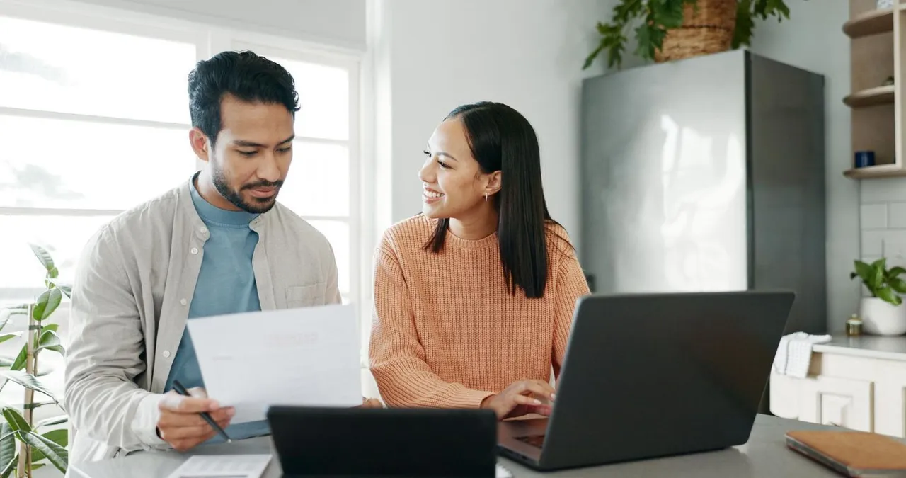 Couple reviewing paperwork and laptop at a table in a well-lit kitchen.