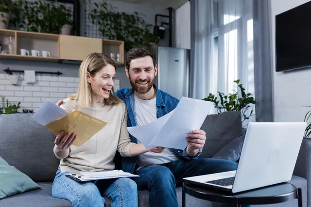 Couple on a sofa reviewing documents with a laptop on the table.
